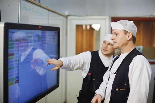 To menn i arbeidstøy og hvite hetter ser på en berøringsskjerm og diskuterer data på skjermen.