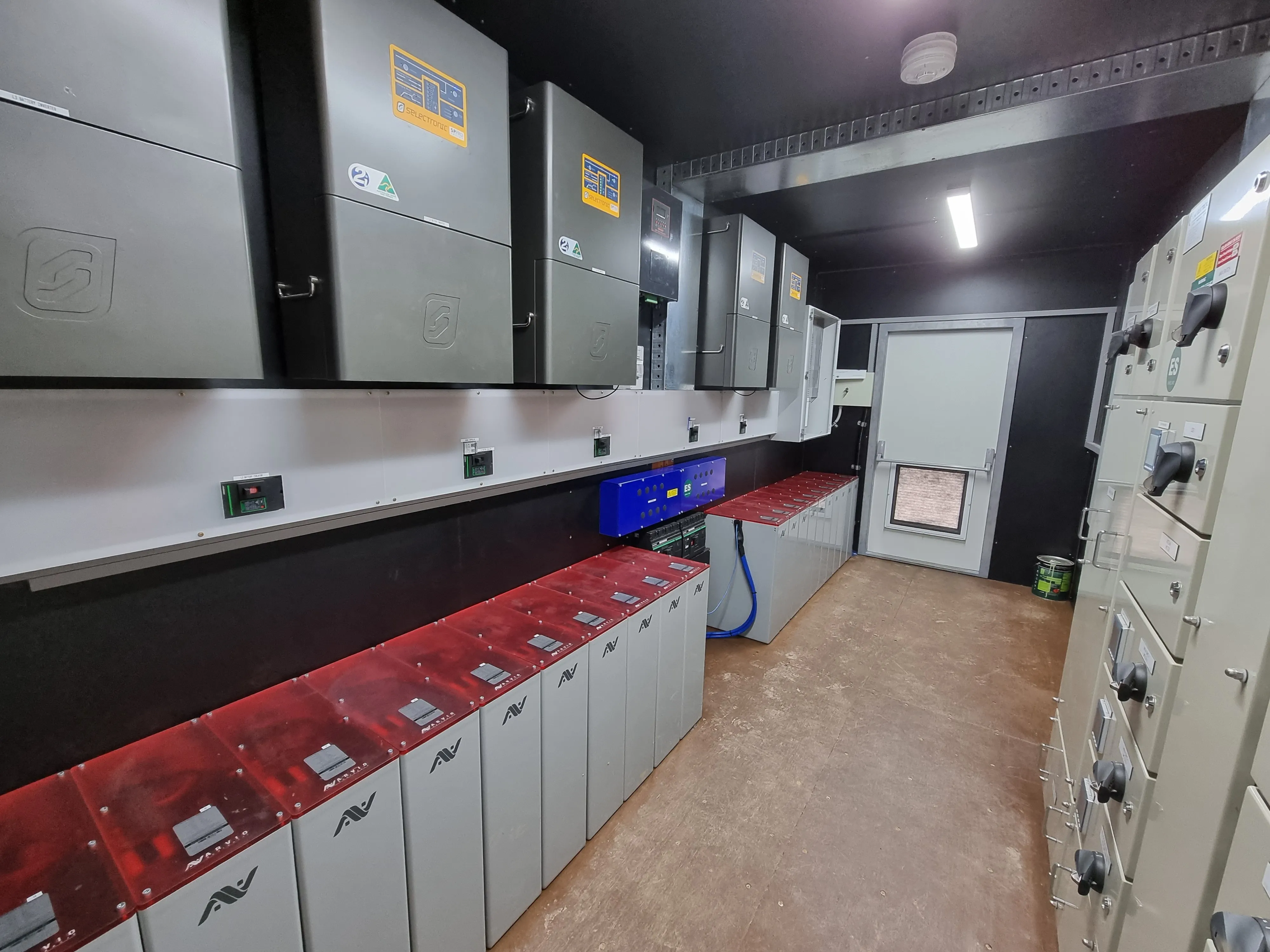 Room with rows of electrical control panels and units along walls under fluorescent lighting.