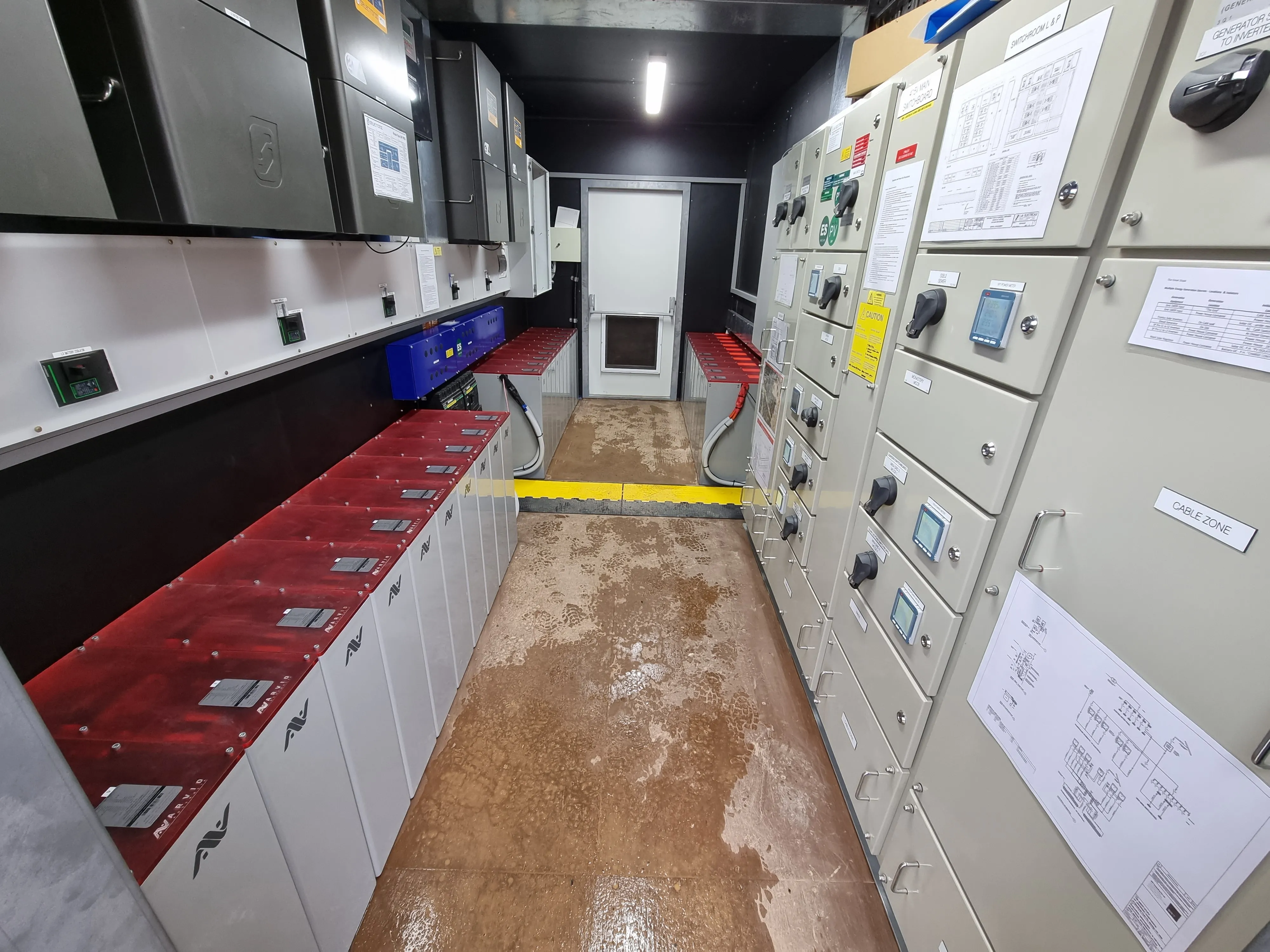 Industrial room with electrical panels, control units, and red-topped battery cabinets along walls.