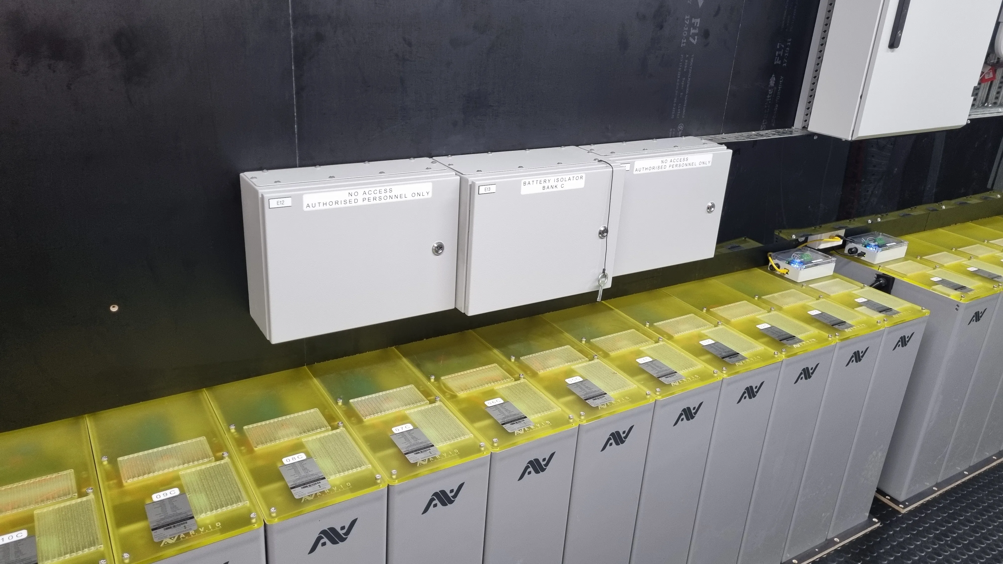 Row of industrial battery units with yellow covers below three grey control boxes on a black wall.
