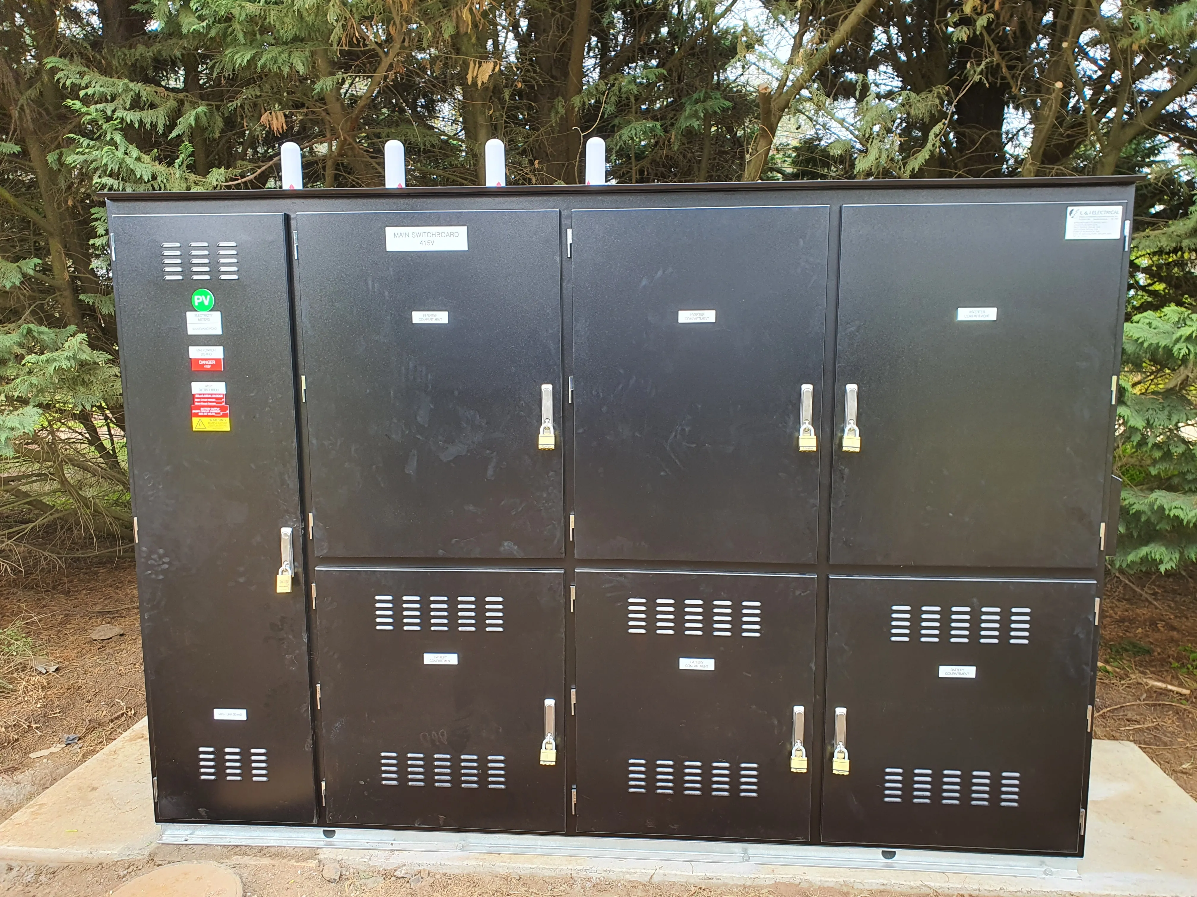 Black electrical switchboard cabinet with locked doors outdoors near trees.