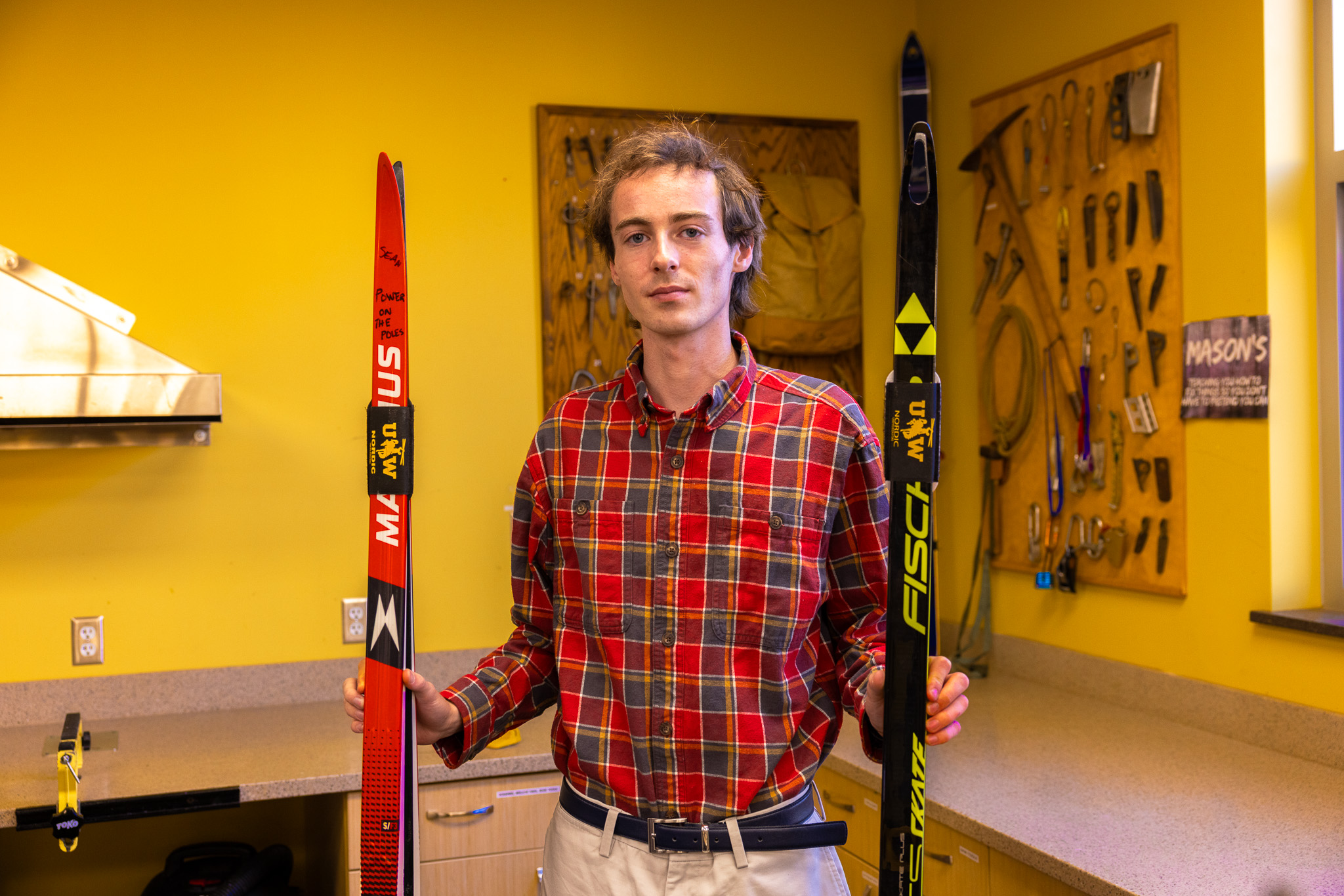 Young man in a red plaid shirt holding a pair of skis inside a room with yellow walls and tools hanging on a board.