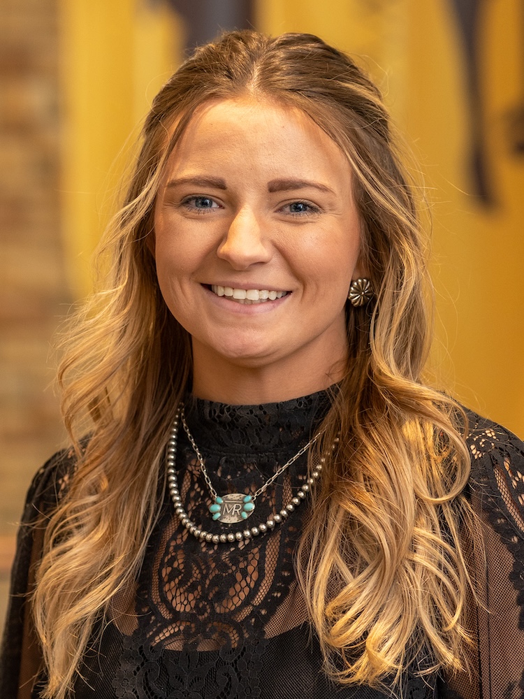 Smiling woman with long wavy blonde hair wearing a black lace top and layered necklaces.