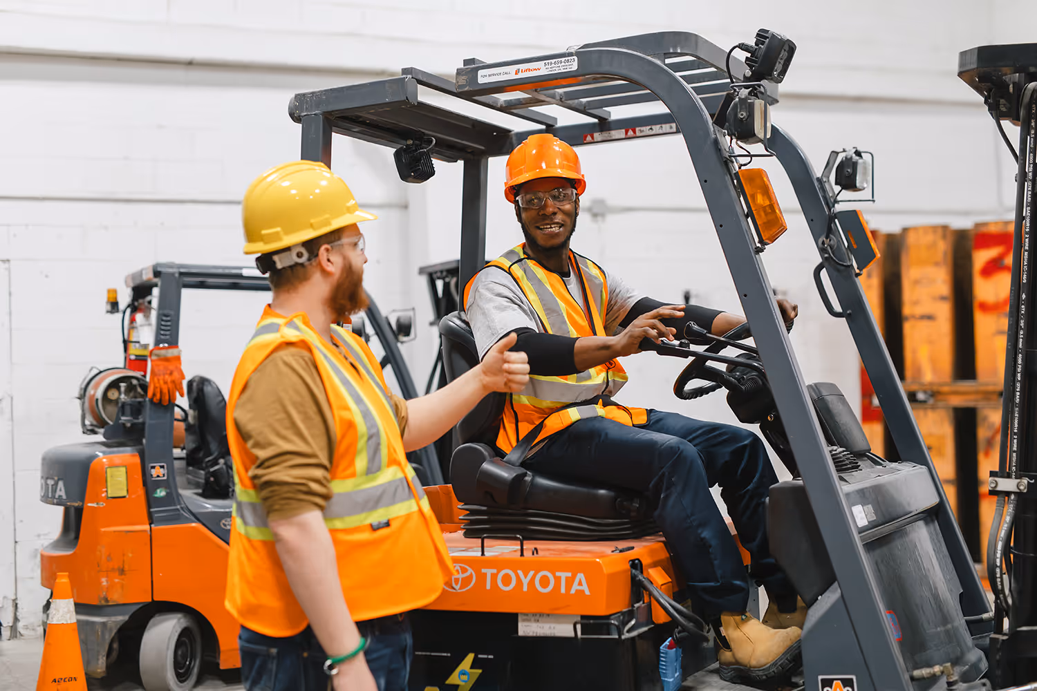 Two Pathways students in hi-vis vests and hard hats, one is operating a forklift while the other looks on.
