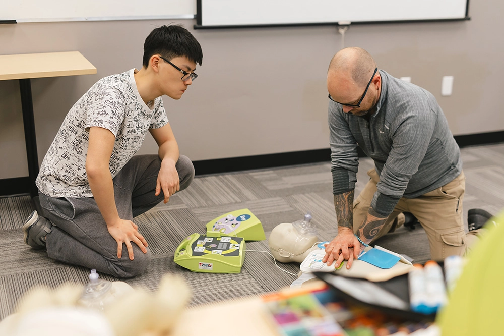 Instructor demonstrating CPR on a CPR training manikin while a Pathways Certification participant looks on