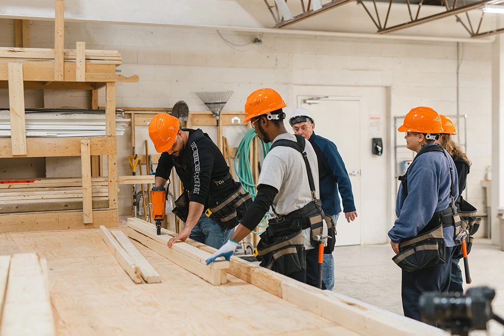 Construction Technology Training participants in hard hats and toolbelts look on as classmate uses drill on piece of wood