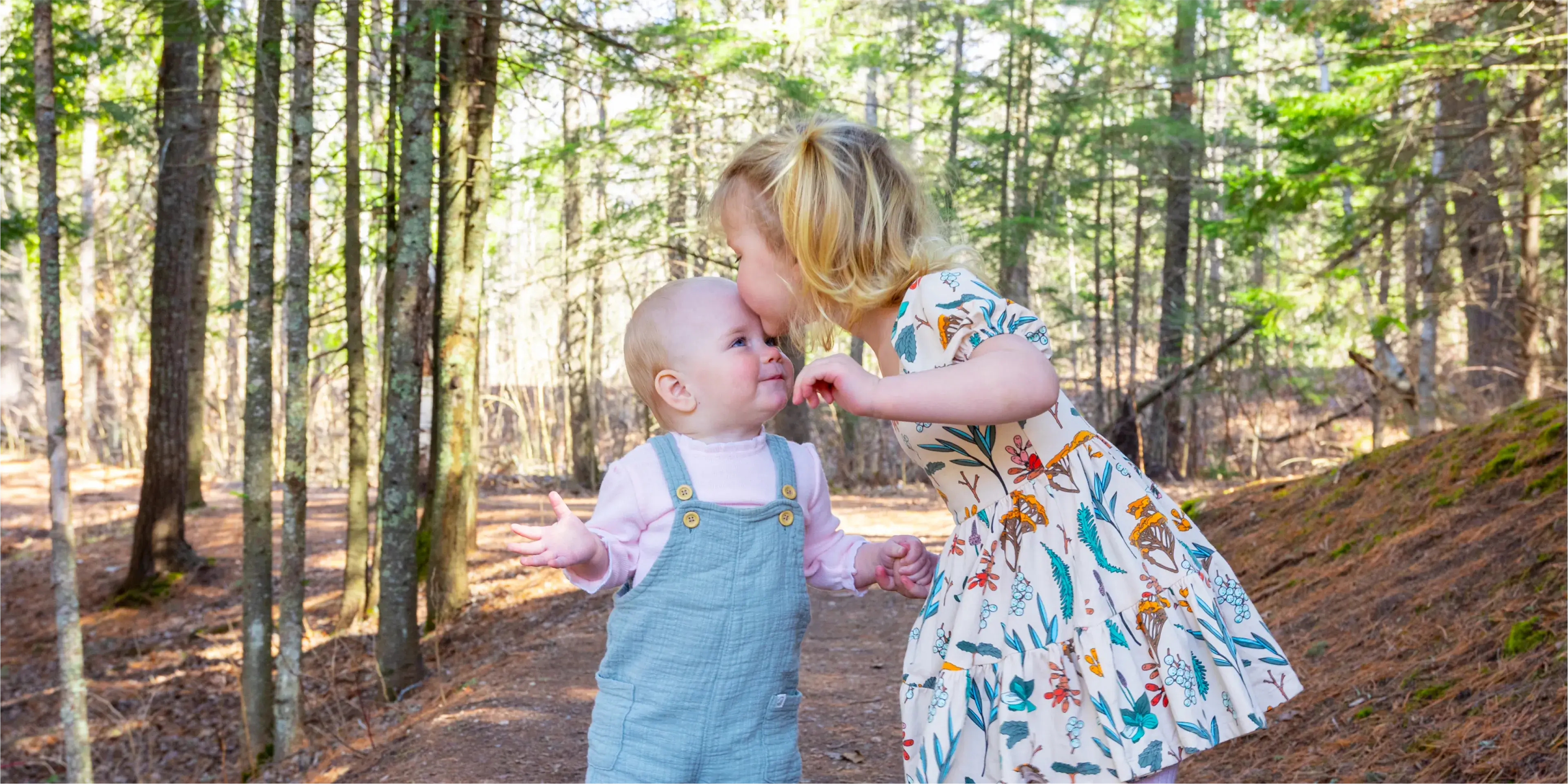 A young girl kisses a baby on the cheek while standing on a forest path.