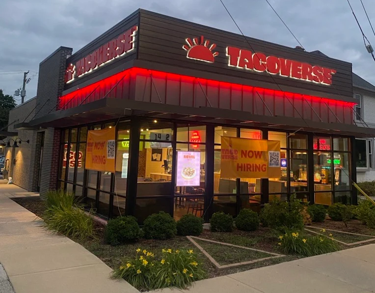 Tacoverse restaurant building at dusk with illuminated red signage and 'Now Hiring' banners on windows.