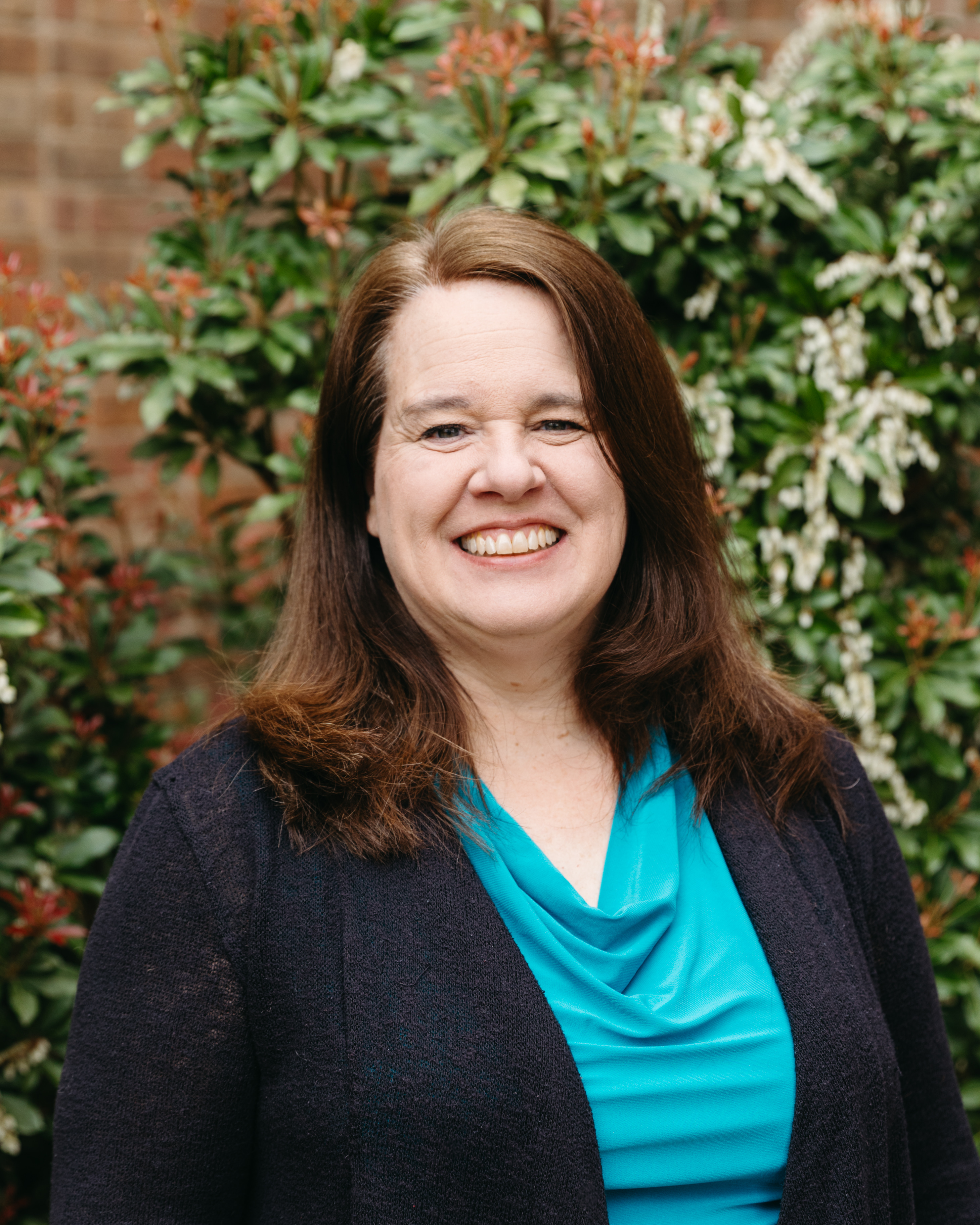 Smiling woman with shoulder-length brown hair wearing a blue top and dark cardigan standing in front of green leafy plants.