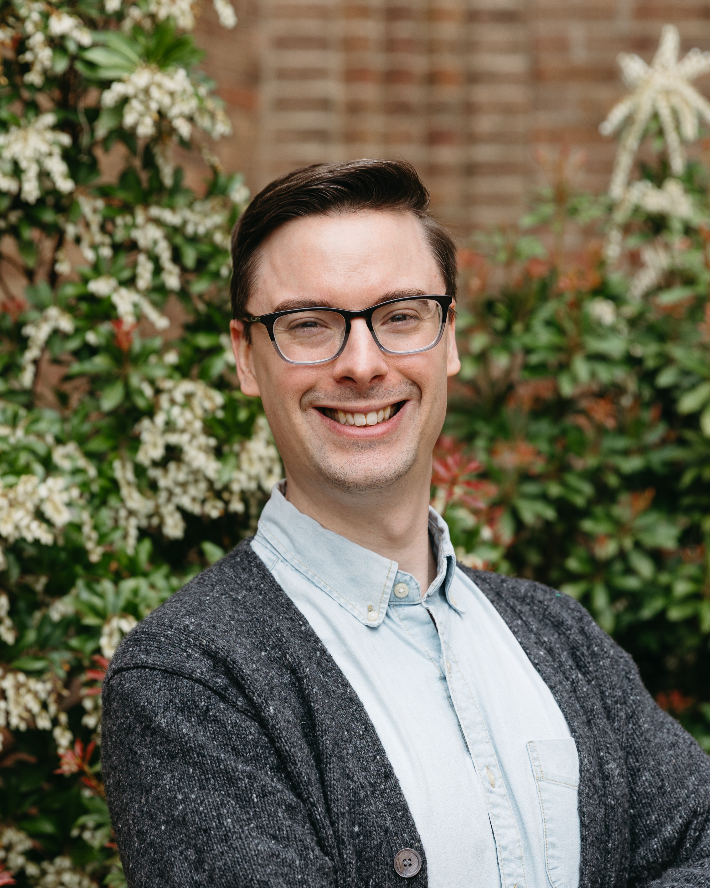 Smiling man with glasses wearing a light blue shirt and dark gray cardigan standing outdoors in front of flowering bushes.