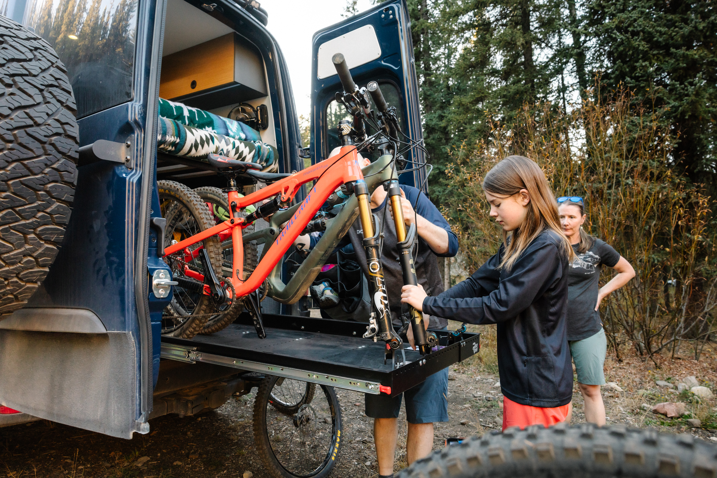 Keira prepares to take her mountain bike out of the van's gear tray, while Frank and Nathalie watch from a distance.=