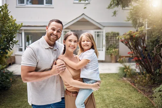 A young family stands happily outside their house, with coverage from an independent insurance agency in South Florida.