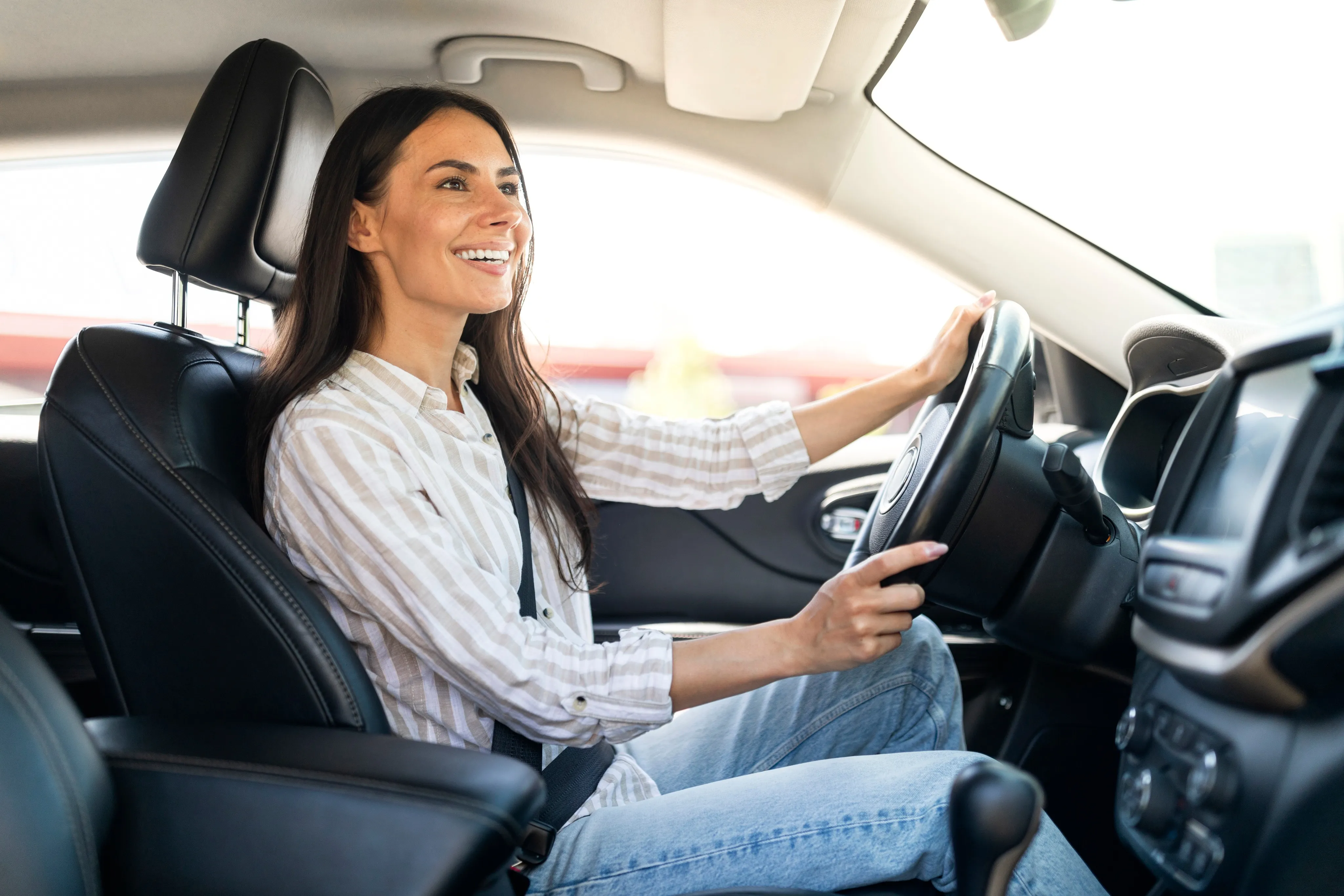 A cheerful woman in a striped blouse and jeans drives her car with a smile, representing the peace of mind that comes from choosing an independent insurance agency in South Florida.