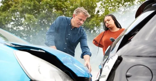 A man and a young woman inspect the damage to their cars after a collision, with smoke rising from the vehicles, showing the value of claims assistance from an independent insurance agency in South Florida.