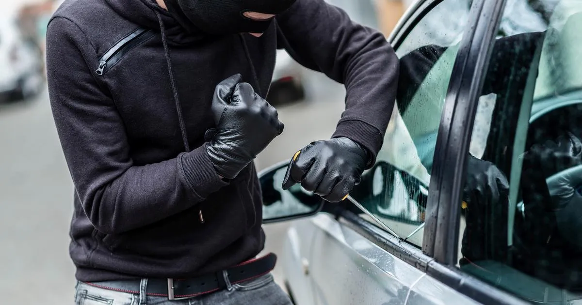 A masked thief in black gloves and a hoodie tries to break into a car, demonstrating the need for theft protection offered by an independent insurance agency in South Florida.