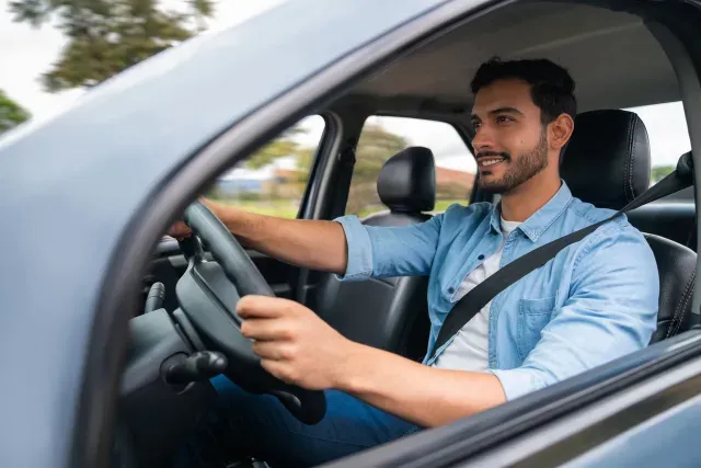 A young man wearing a blue shirt and seatbelt drives his car on a clear day, showing how everyday drivers rely on the protection of an independent insurance agency in South Florida.