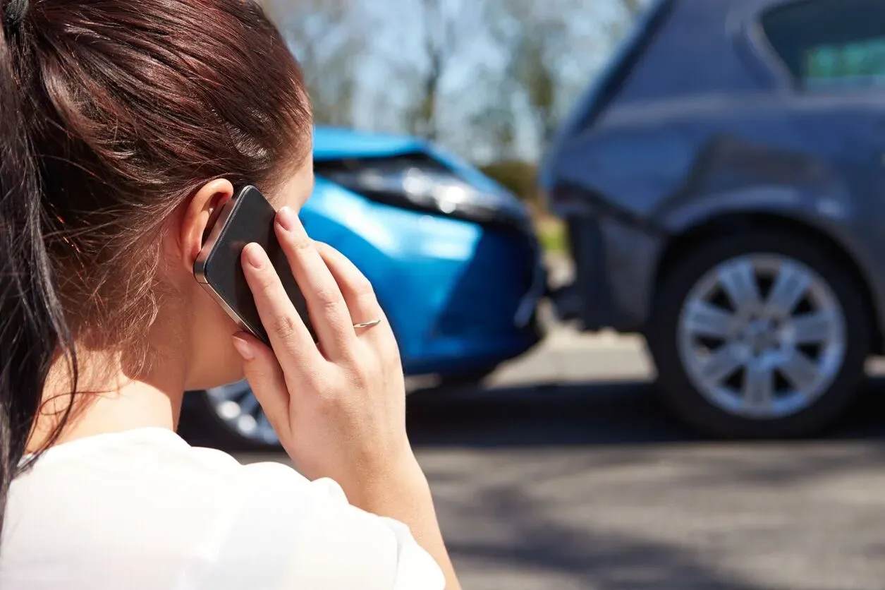 A woman with a ponytail calls for help on her phone after a rear-end accident, with two damaged cars in the background, illustrating the fast claims support from an independent insurance agency in South Florida.