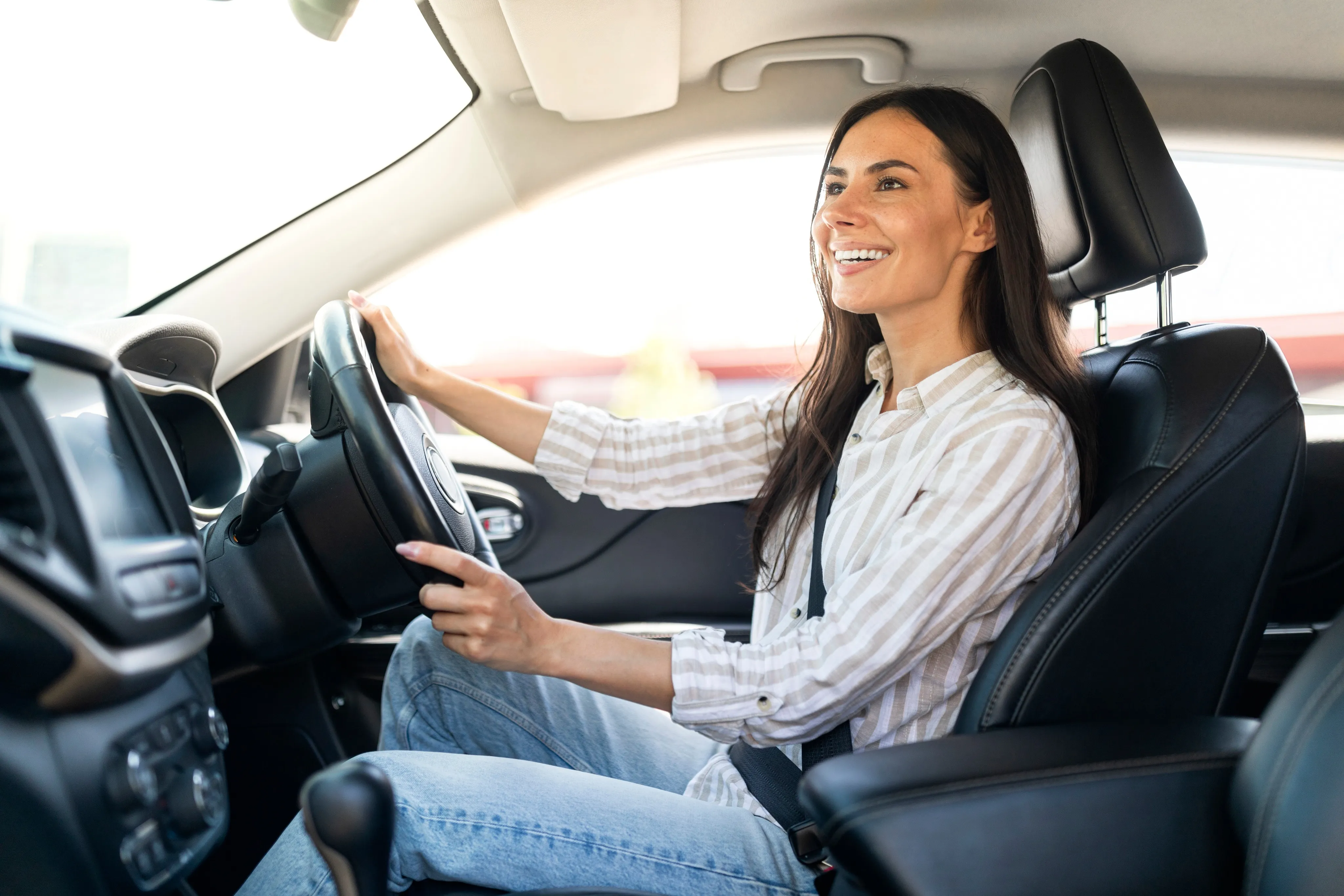 A cheerful woman in a striped blouse and jeans drives her car with a smile, representing the peace of mind that comes from choosing an independent insurance agency in South Florida.