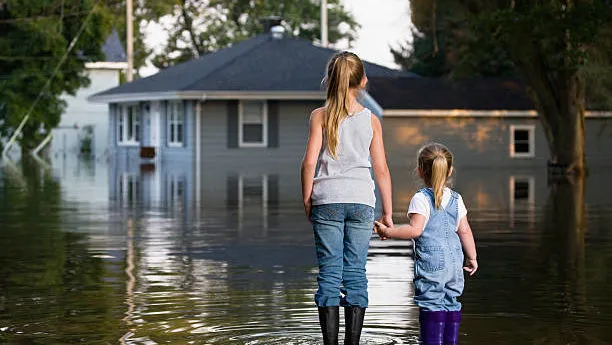 Two young girls in rubber boots hold hands while standing in deep floodwater outside their home, demonstrating the importance of having flood insurance coverage through an independent insurance agency in South Florida.