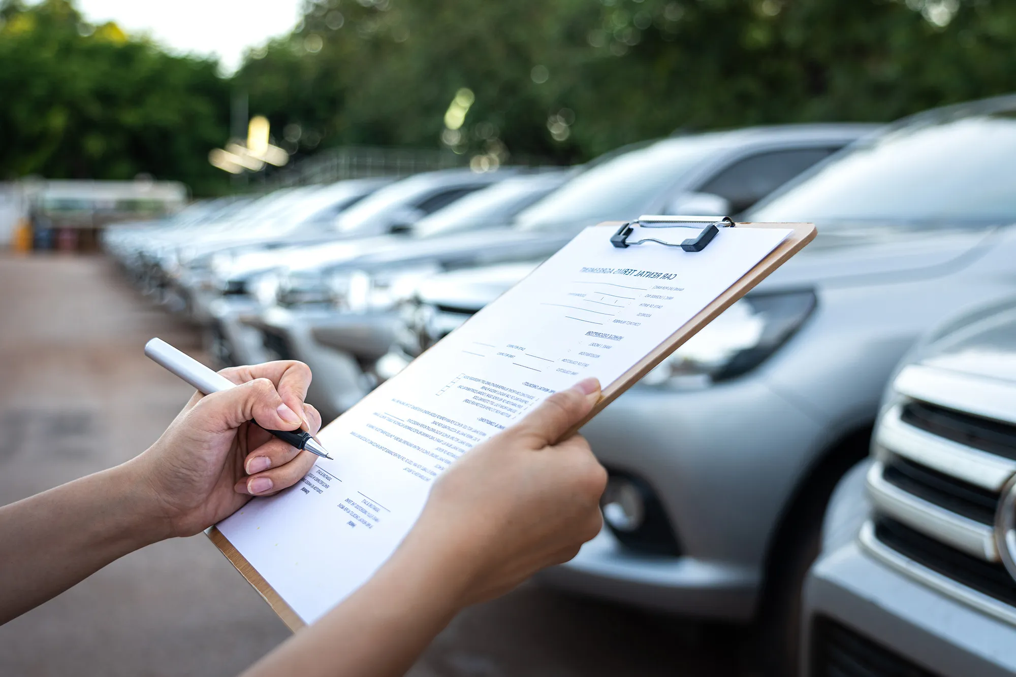Person holding a clipboard and pen with parked cars, ideal for commercial auto insurance.