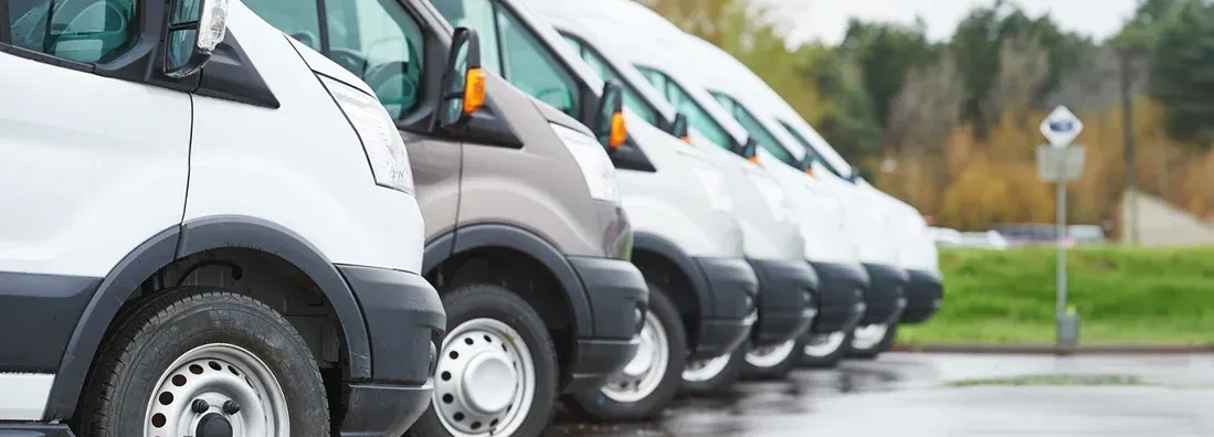 A row of parked white and gray delivery vans on a wet pavement, with trees in background.