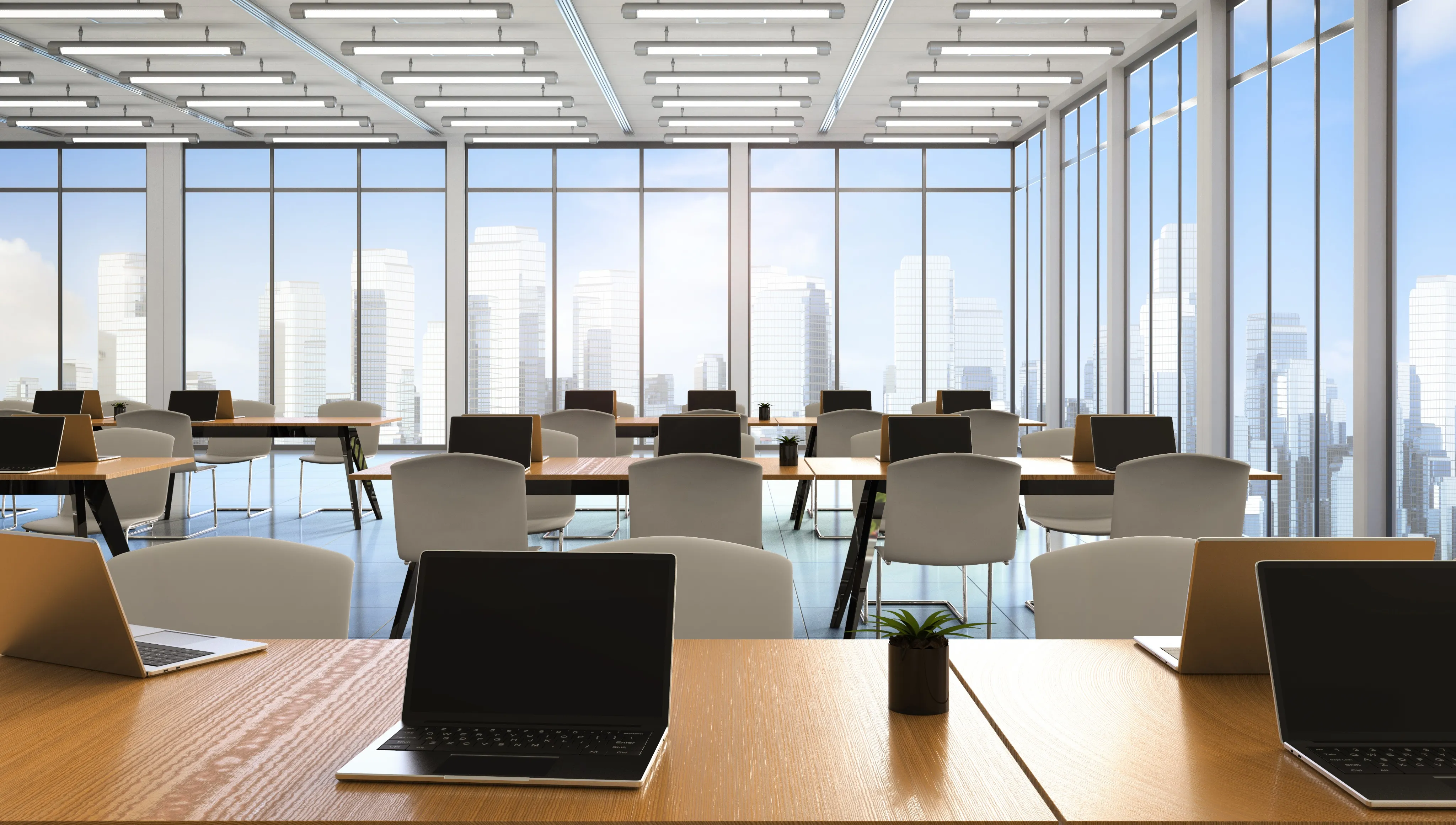Modern office with empty desks, laptops, and large windows overlooking a city skyline.