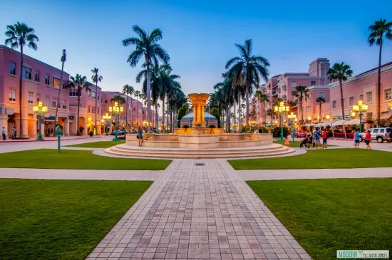 A scenic view of Mizner Park in Boca Raton at dusk, featuring a central fountain, palm trees, and pink buildings.