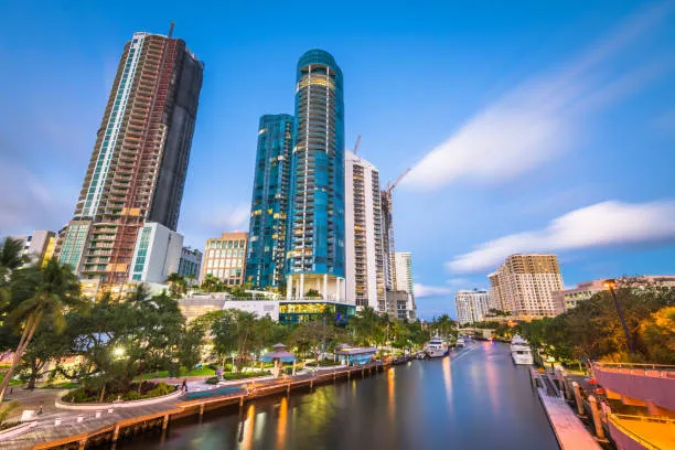 Fort Lauderdale cityscape at dusk with modern high-rise buildings and the riverwalk lined with boats and palm trees.