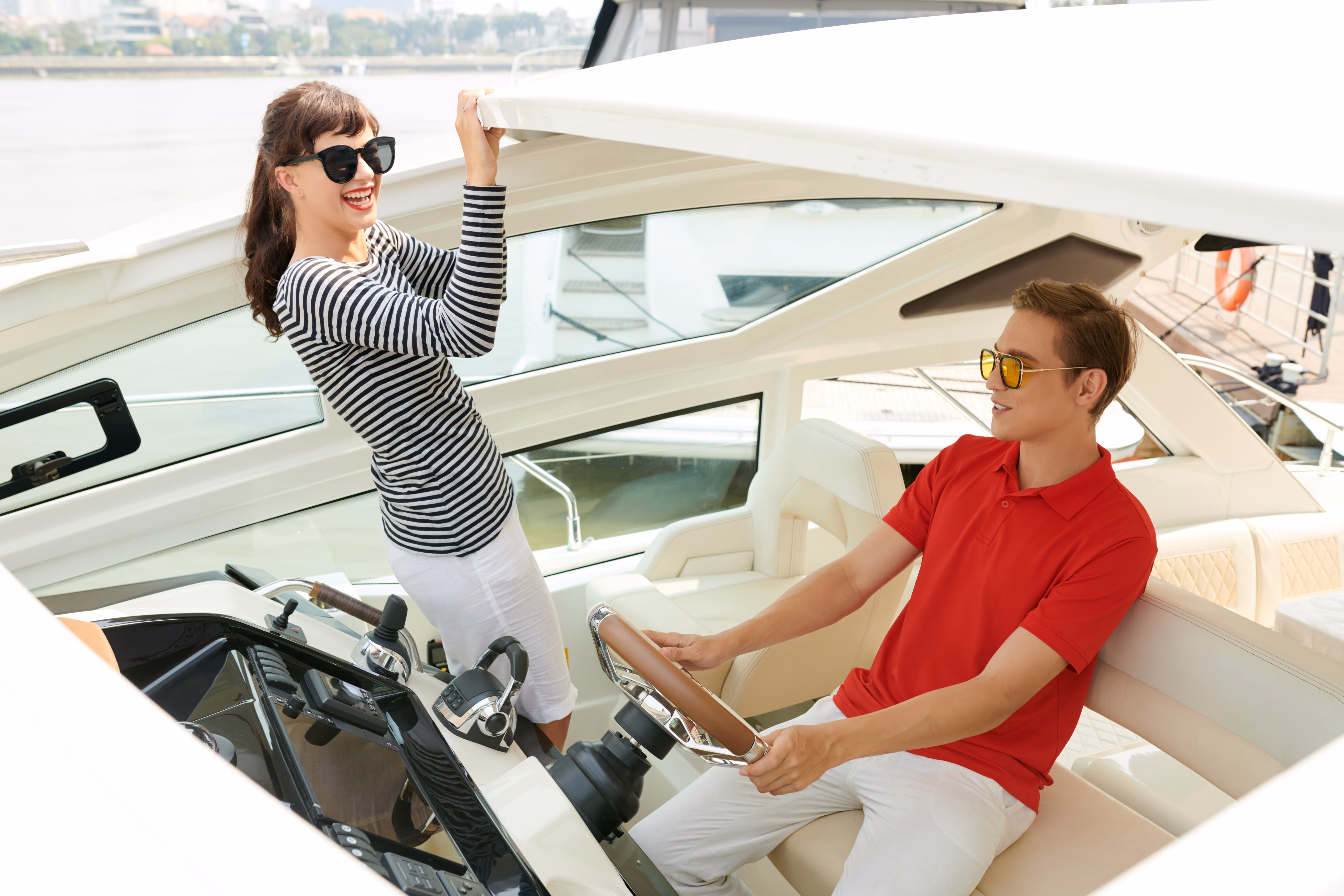 Young woman in striped shirt and sunglasses holding boat roof, smiling at young man in red shirt steering the boat.