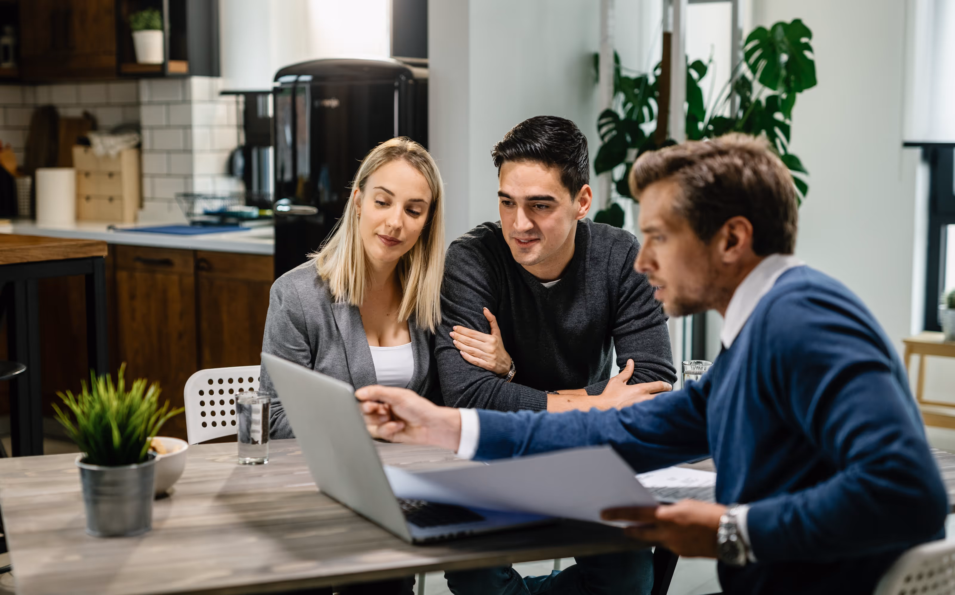 Young couple and real estate agent reviewing housing plans on a laptop in a modern kitchen.