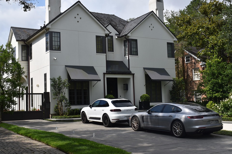 Modern two-story white house with black window awnings and two luxury cars parked in the driveway.