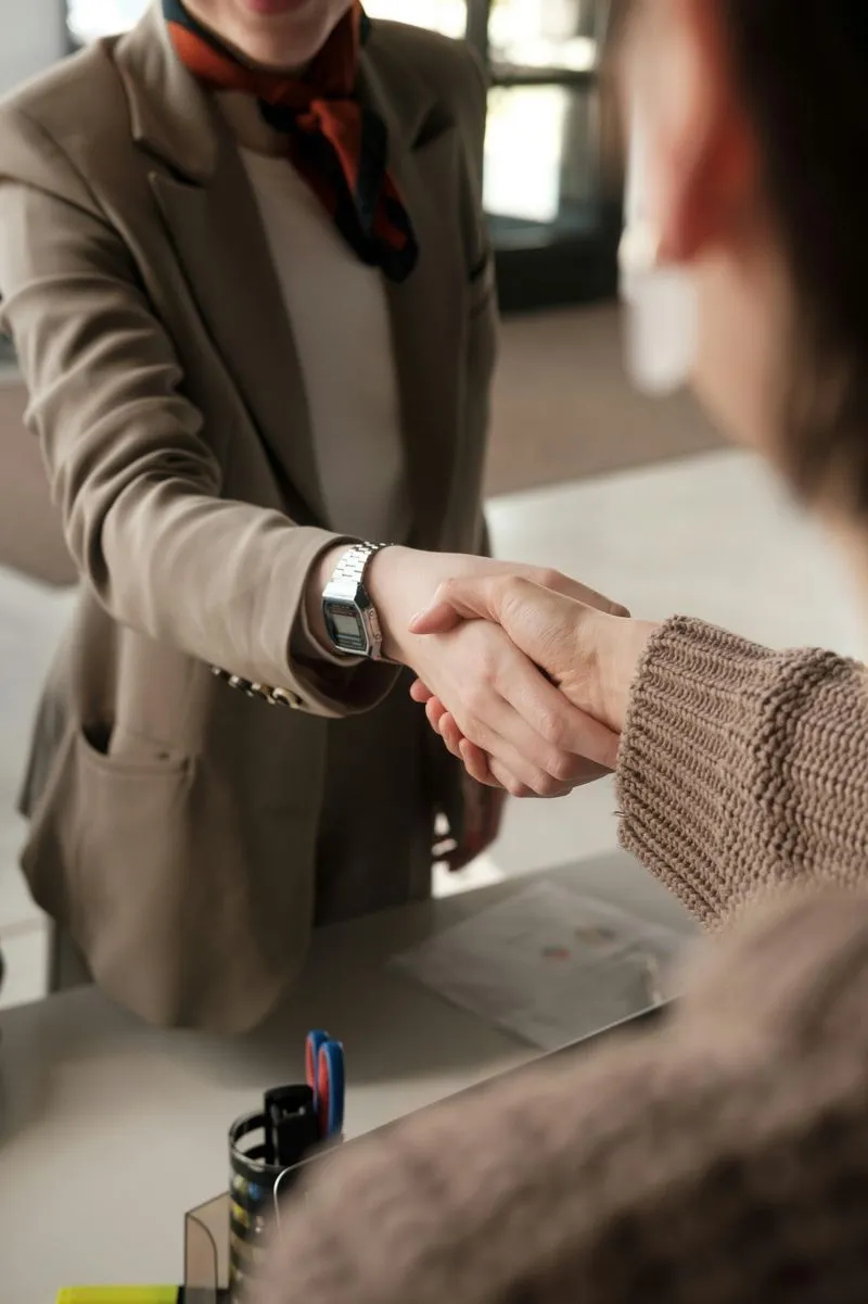 Two people shaking hands across a desk, one wearing a beige blazer and watch, the other a brown knitted sweater.
