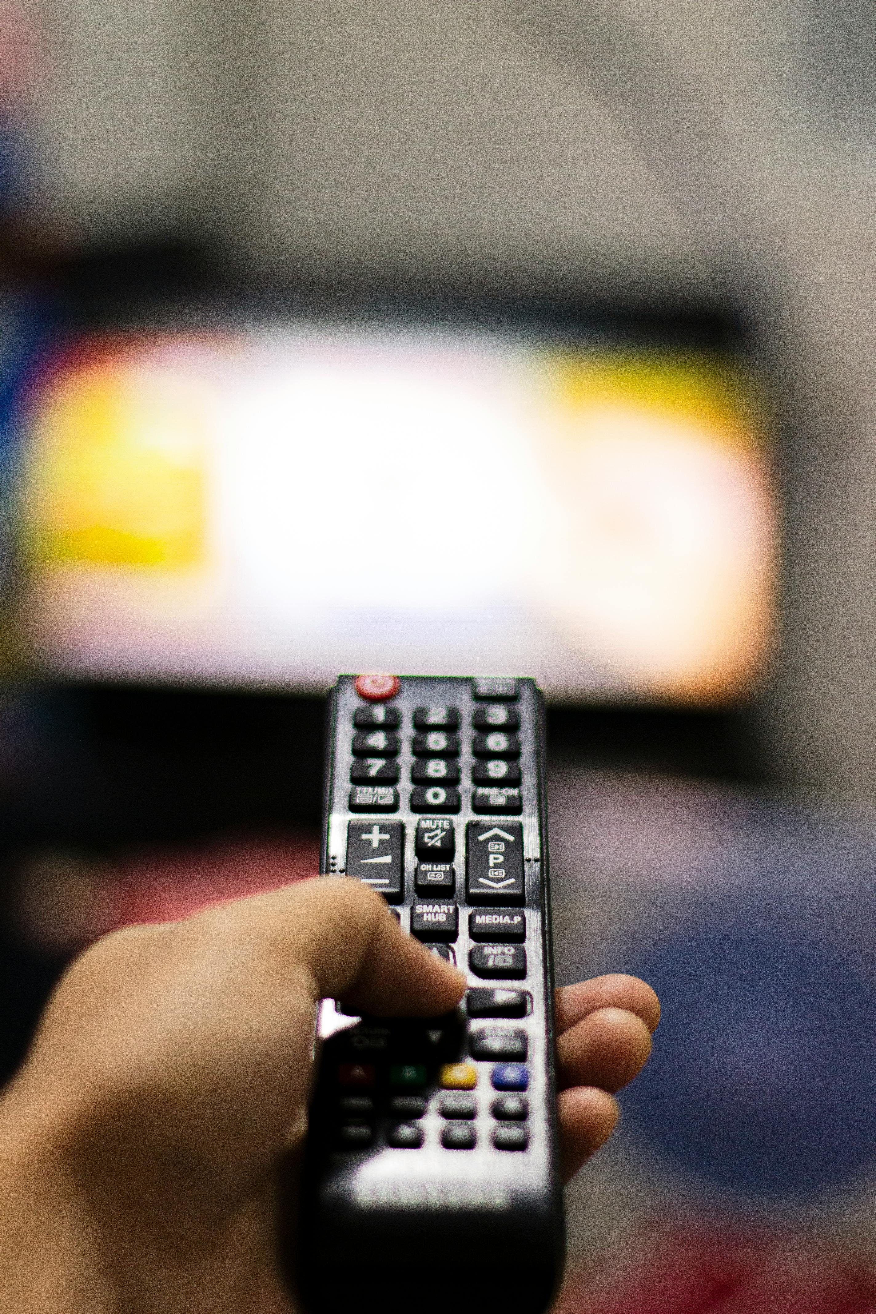 Hand holding a Samsung TV remote pointed at a blurred television screen.