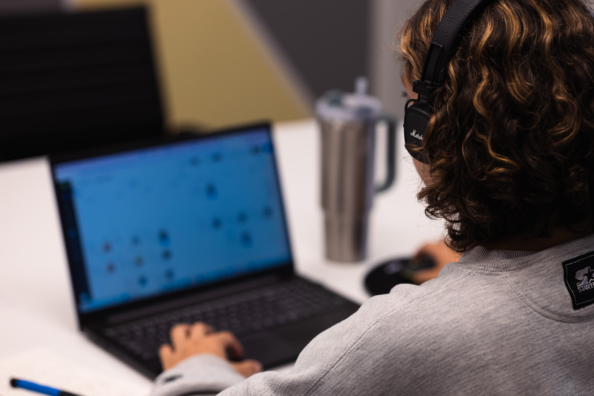 Person wearing headphones and a gray sweatshirt using a laptop at a white desk with a stainless steel tumbler nearby.