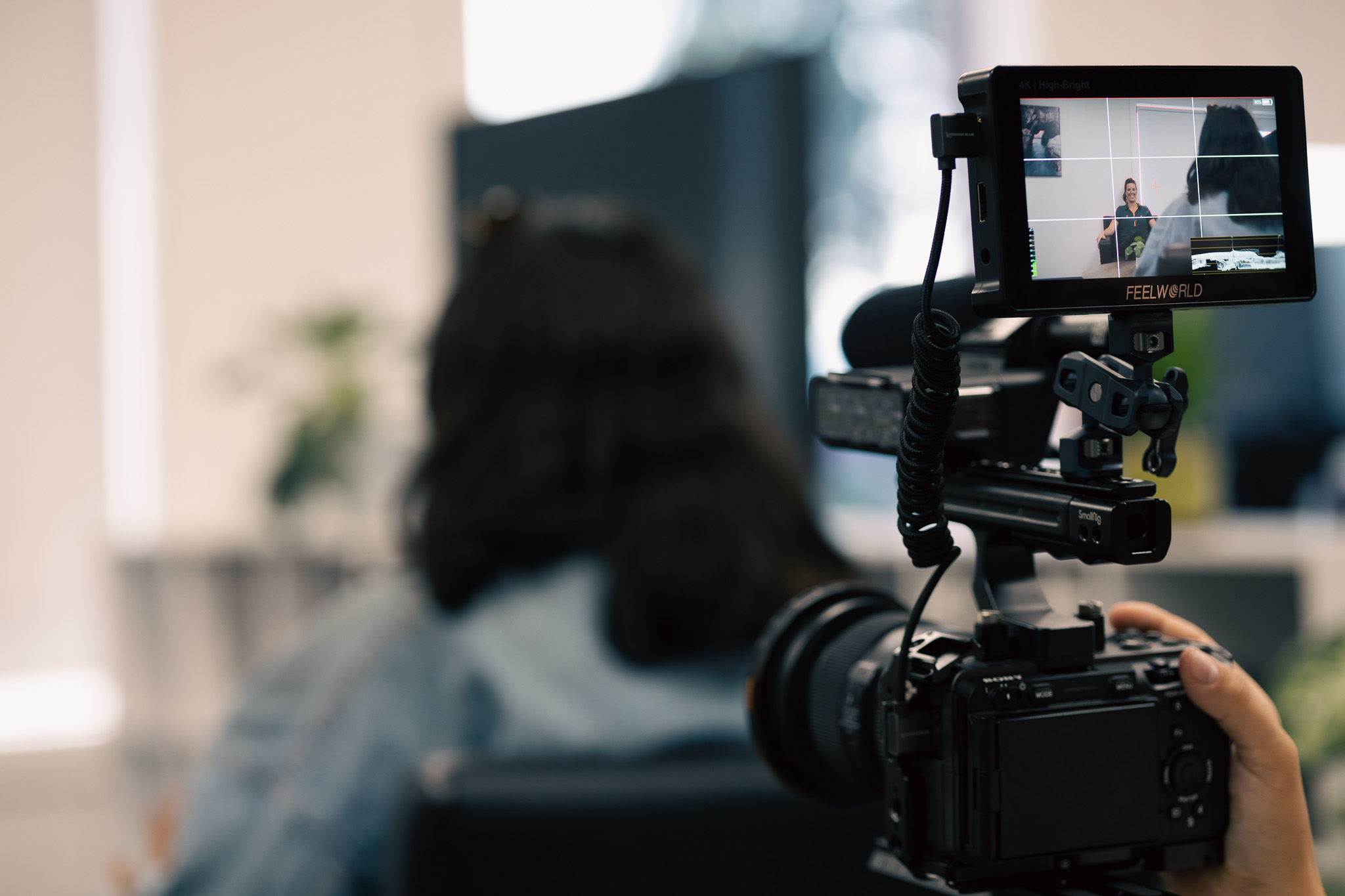Close-up of a professional video camera filming a woman sitting on a chair indoors.