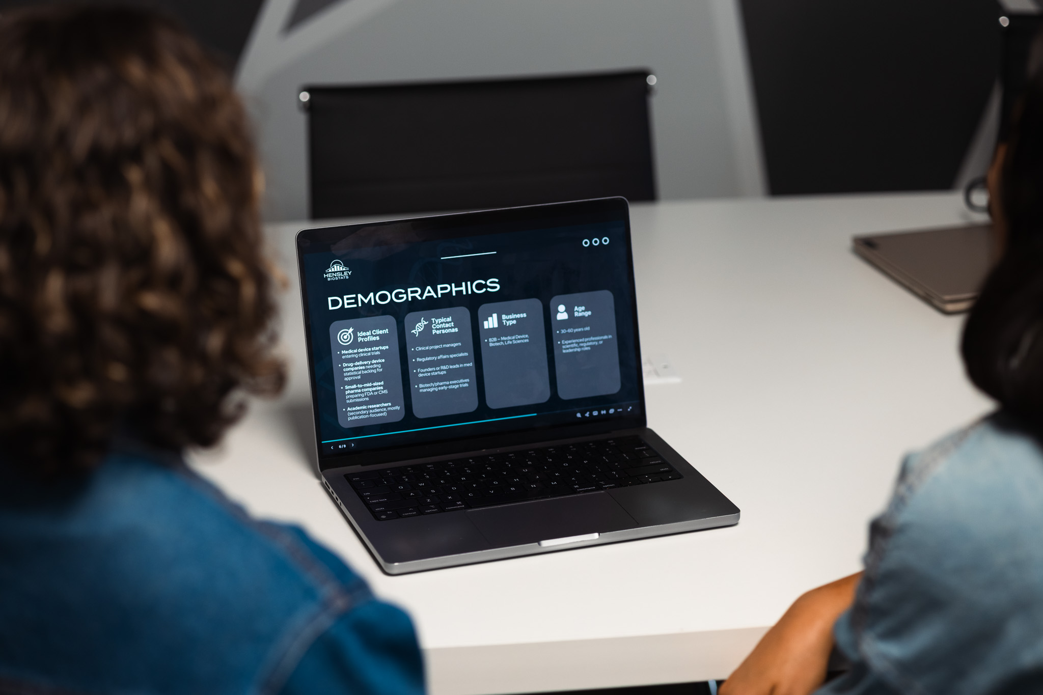 Two people sitting at a table looking at a laptop screen displaying a demographics presentation slide.
