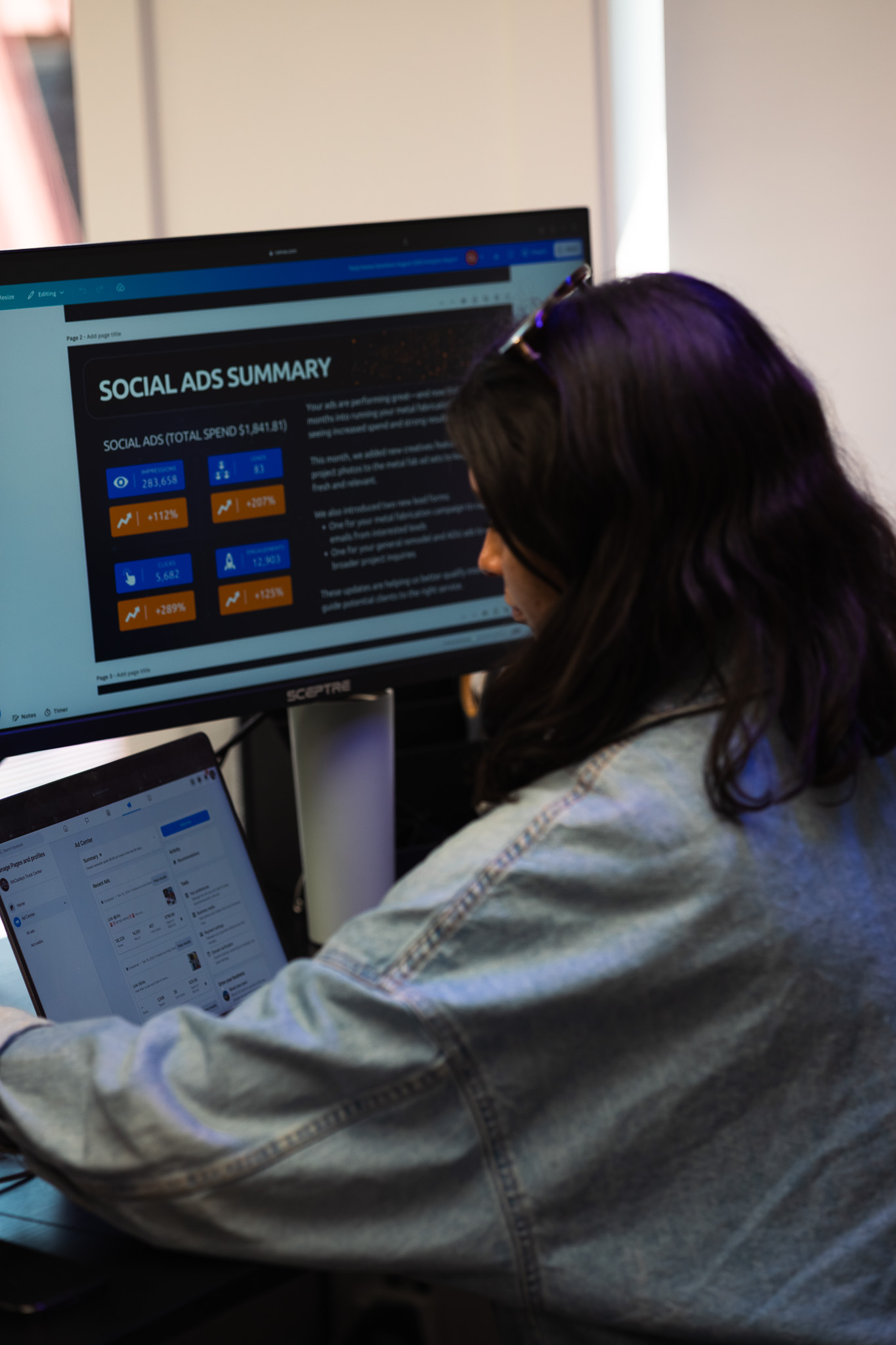 Person in a denim jacket working at a desk with a laptop and a large monitor displaying a social ads summary report.