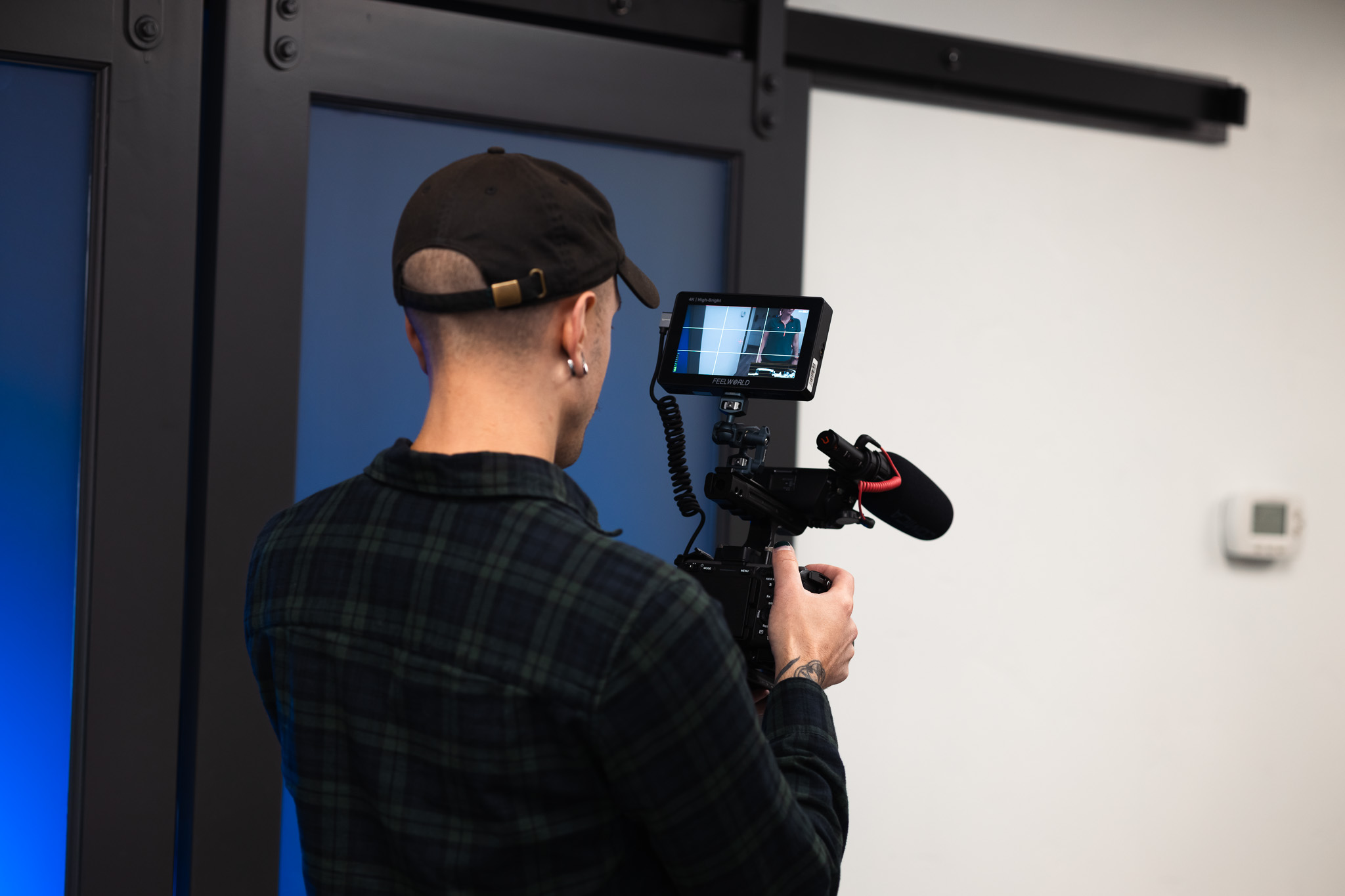 Person wearing a black cap and plaid shirt filming with a professional camera equipped with a microphone and monitor screen.