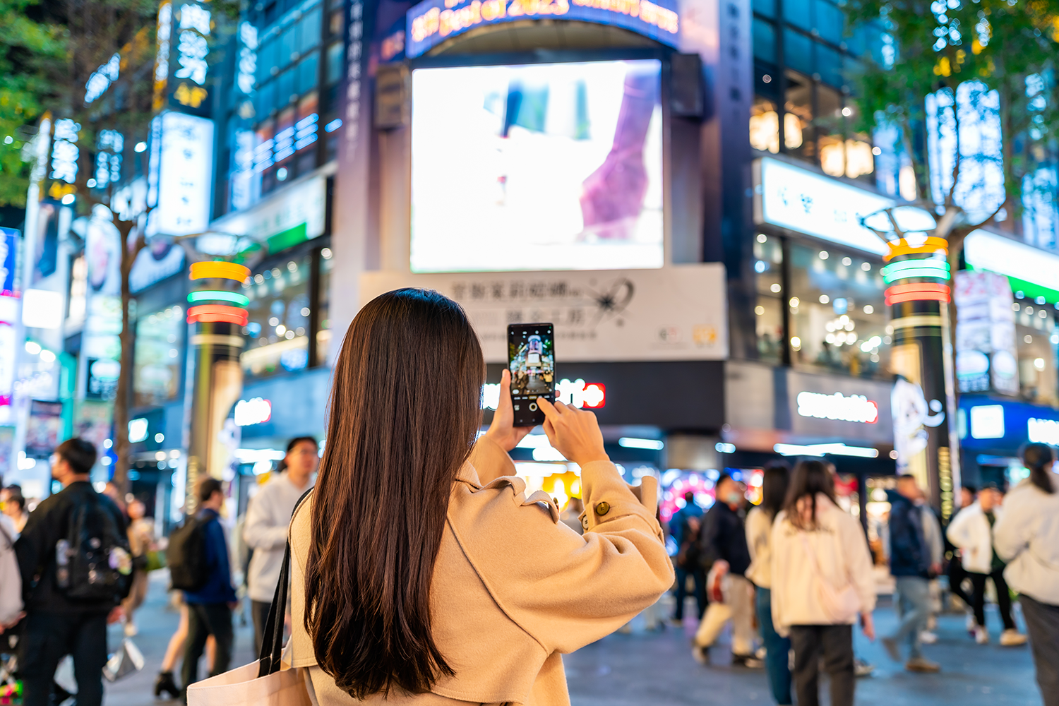 Woman in beige coat taking a photo with smartphone of brightly lit storefronts and billboards in a busy city street at night.