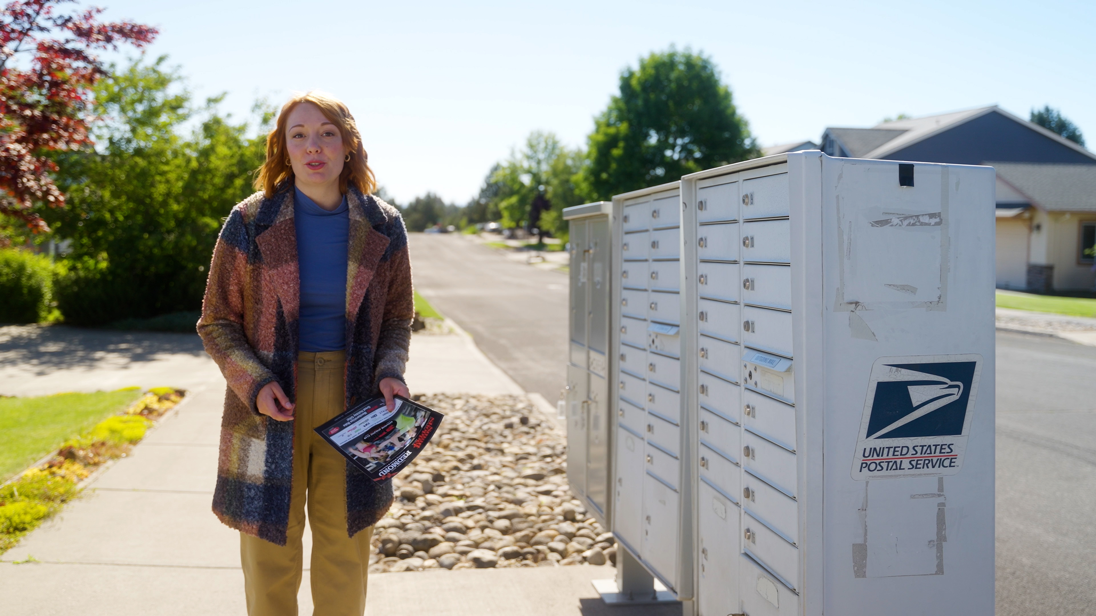 Woman standing on a sidewalk holding flyers next to a United States Postal Service mailbox.