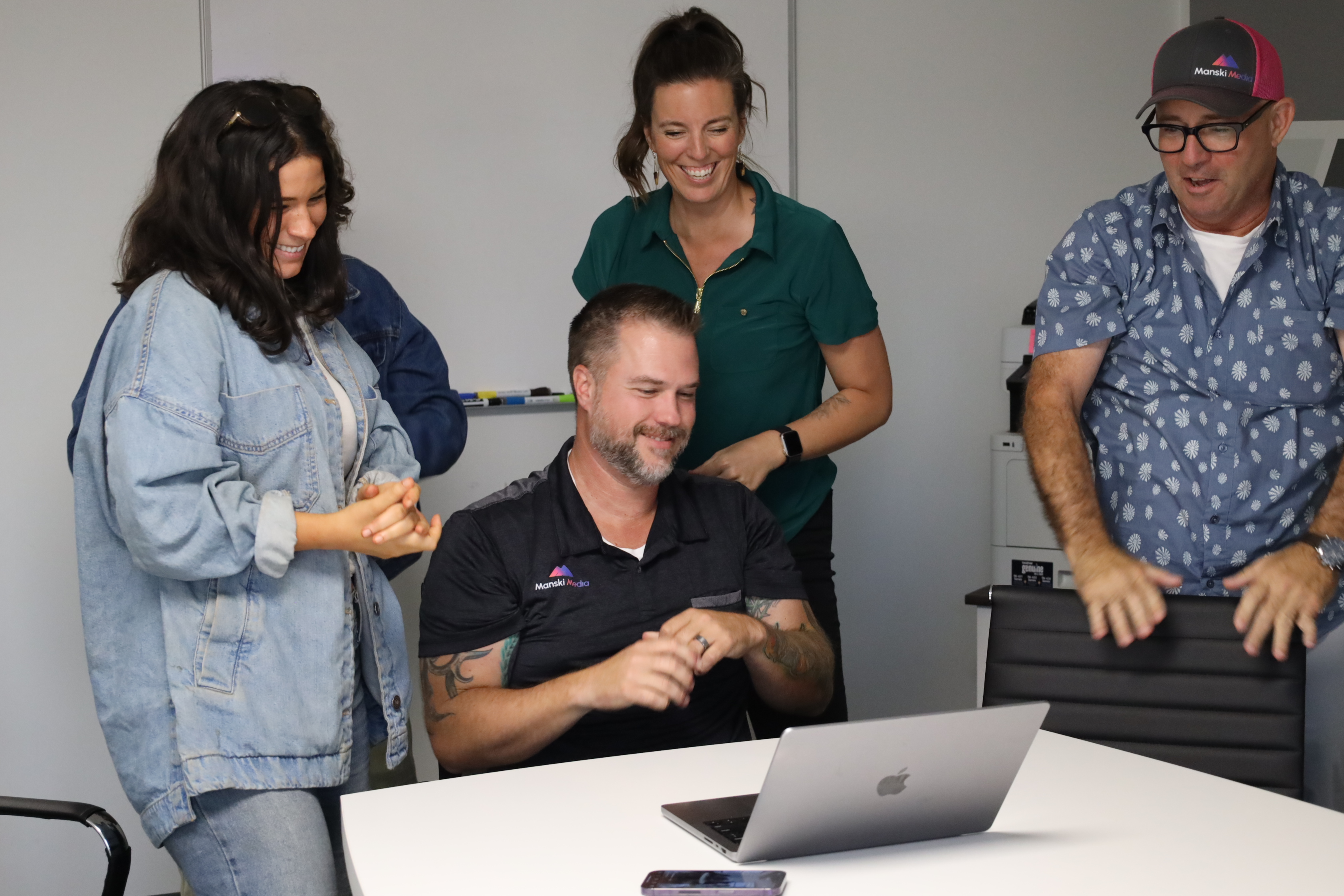 Four people smiling and gathered around a table with a laptop, engaged in a cheerful discussion.
