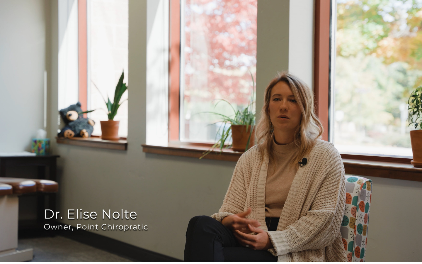 Woman in a beige sweater sitting in a colorful chair near windows with plants, speaking.