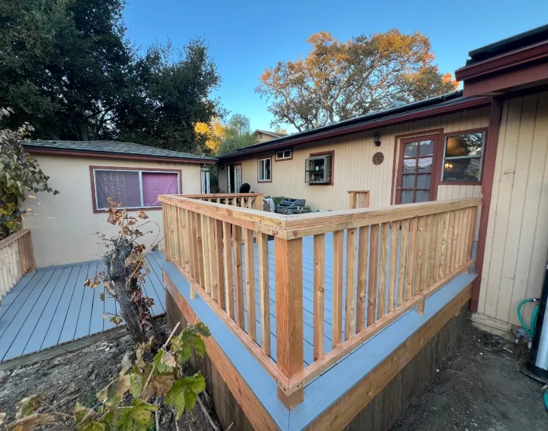 Newly built wooden deck with railing attached to a single-story beige house, surrounded by trees