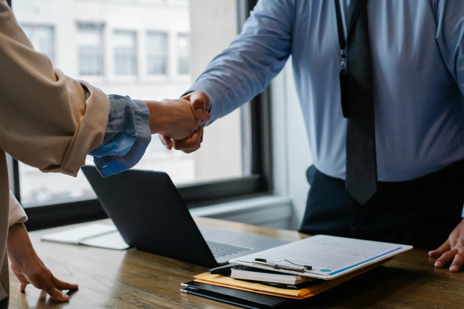 Two individuals shaking hands over a desk with office supplies.