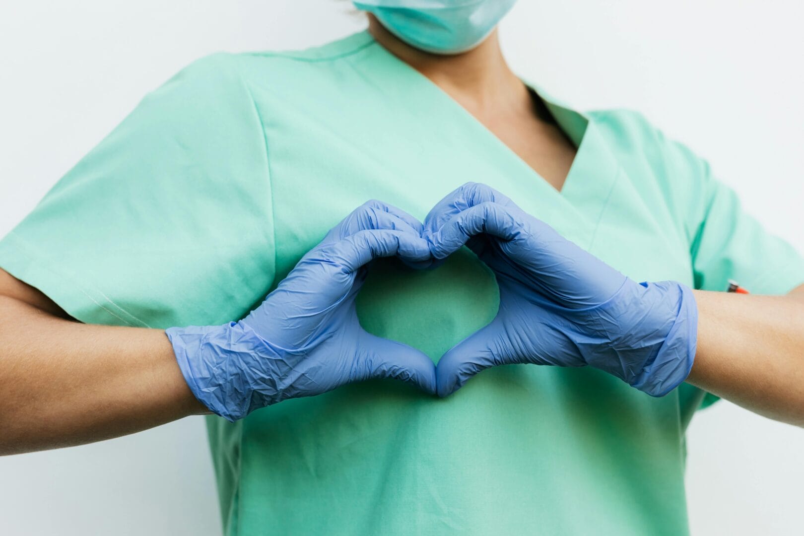 Person wearing scrubs and medical gloves forming a heart shape with their hands.