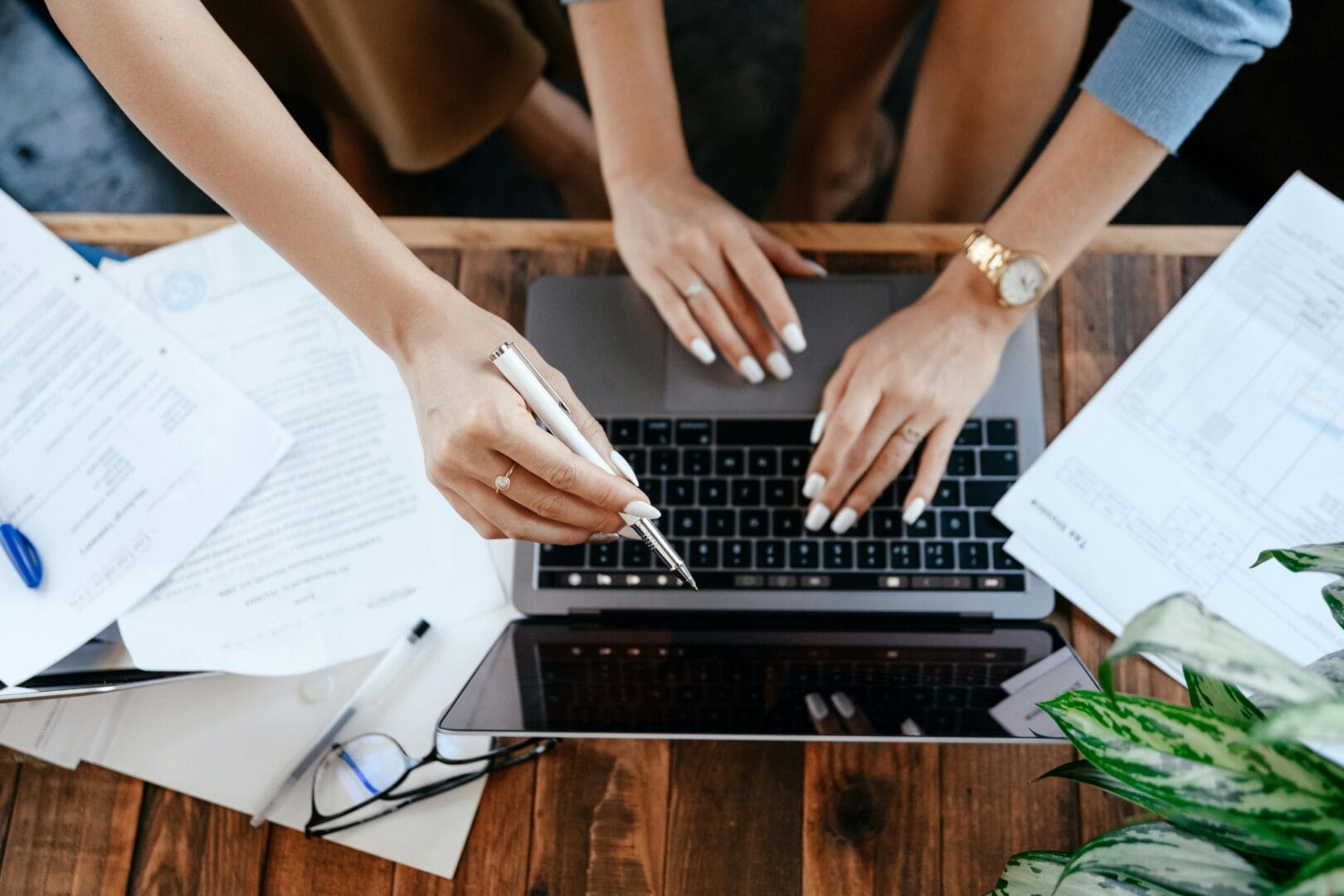 Two people collaborating over a laptop with papers and a plant on a wooden table.