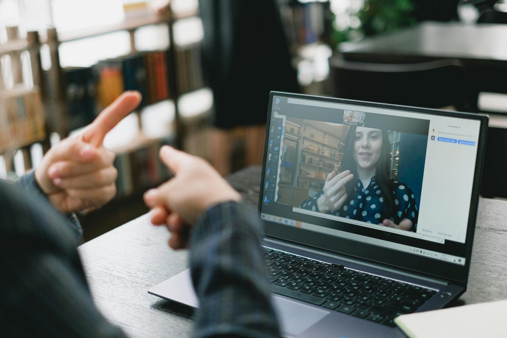 Person in a video call on a laptop, with another person visible gesturing in front of the screen.