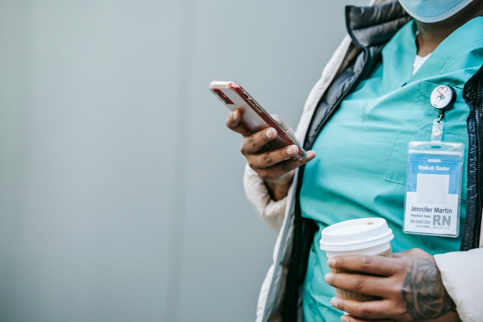 Person in medical scrubs holding a smartphone and a coffee cup.
