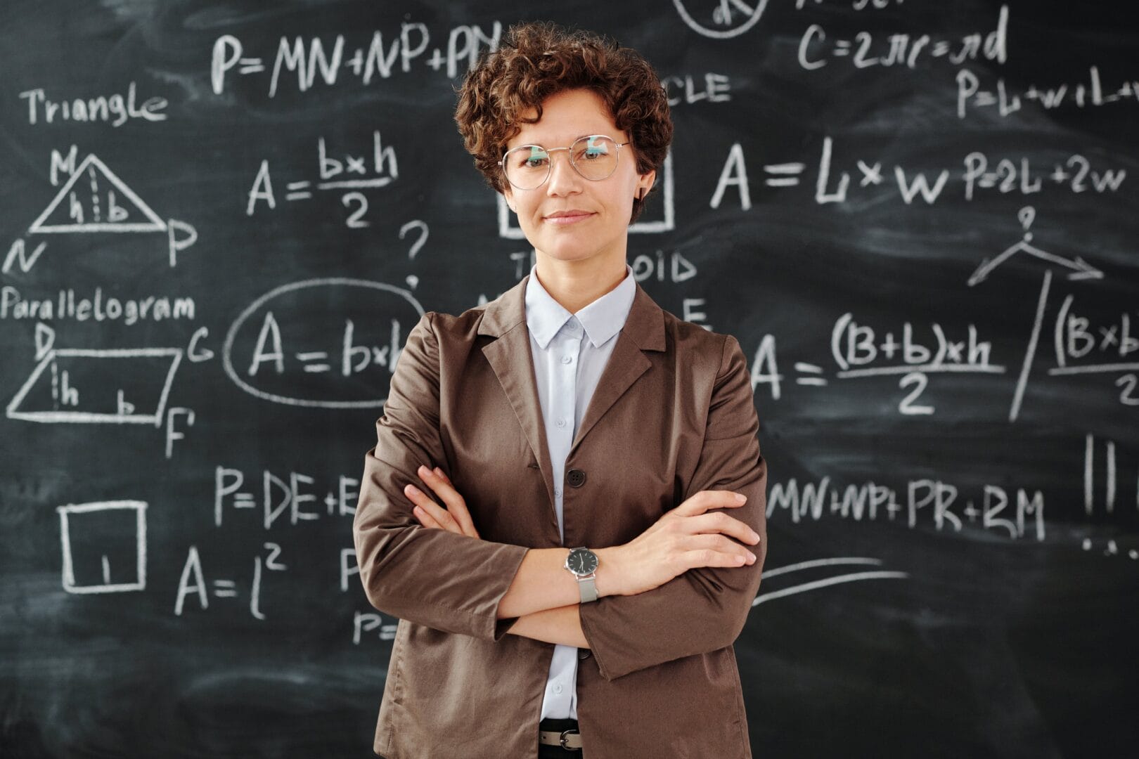 Person with glasses and a brown blazer standing in front of a chalkboard filled with mathematical equations.