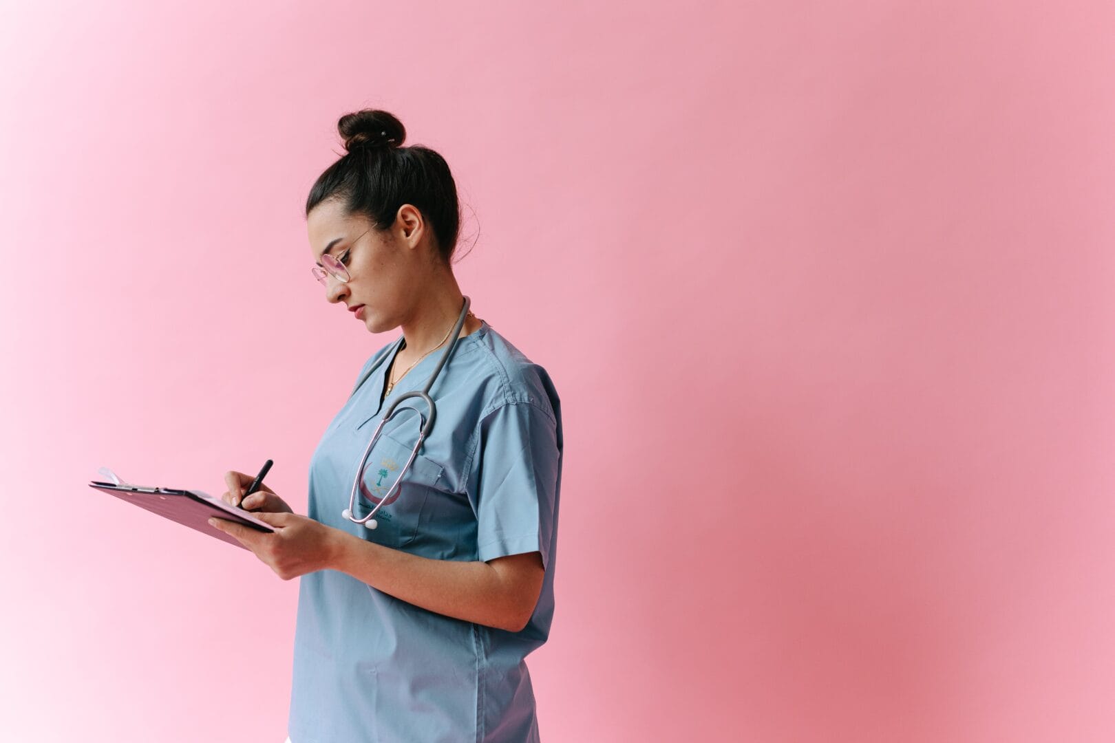 Woman in medical scrubs writing on a clipboard against a pink background.